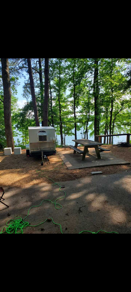 An RV rental camper trailer is set up at a serene lakeside campsite with a picnic table, perfect for nature getaways.