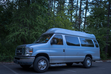 Silver high-roof Ford van, perfect for RV rental adventures, parked amidst tall trees in a lot.