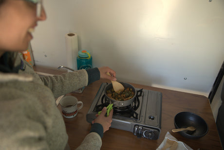 RV rental kitchen: A person cooks a meal on a portable gas stove, with a "Happy Camper" mug beside them.