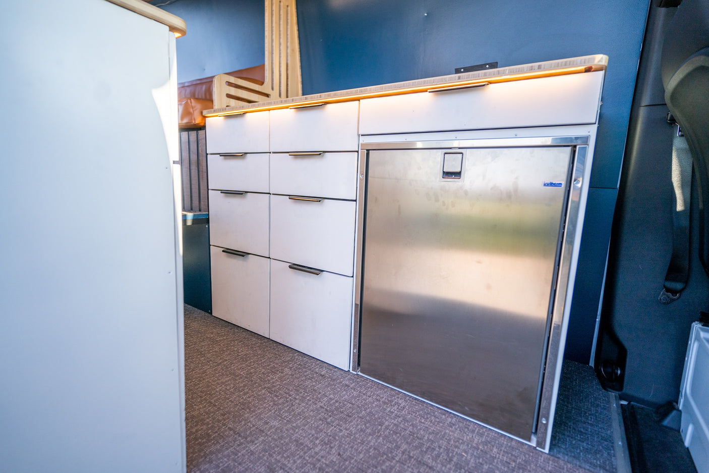 Organized RV rental interior: modern white drawers, integrated stainless steel fridge, and warm LED countertop lighting.