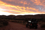 A dark RV rental Jeep with rooftop tent on a dirt road, facing dark mountains under a vibrant orange sunset sky.