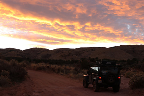 A dark RV rental Jeep with rooftop tent on a dirt road, facing dark mountains under a vibrant orange sunset sky.