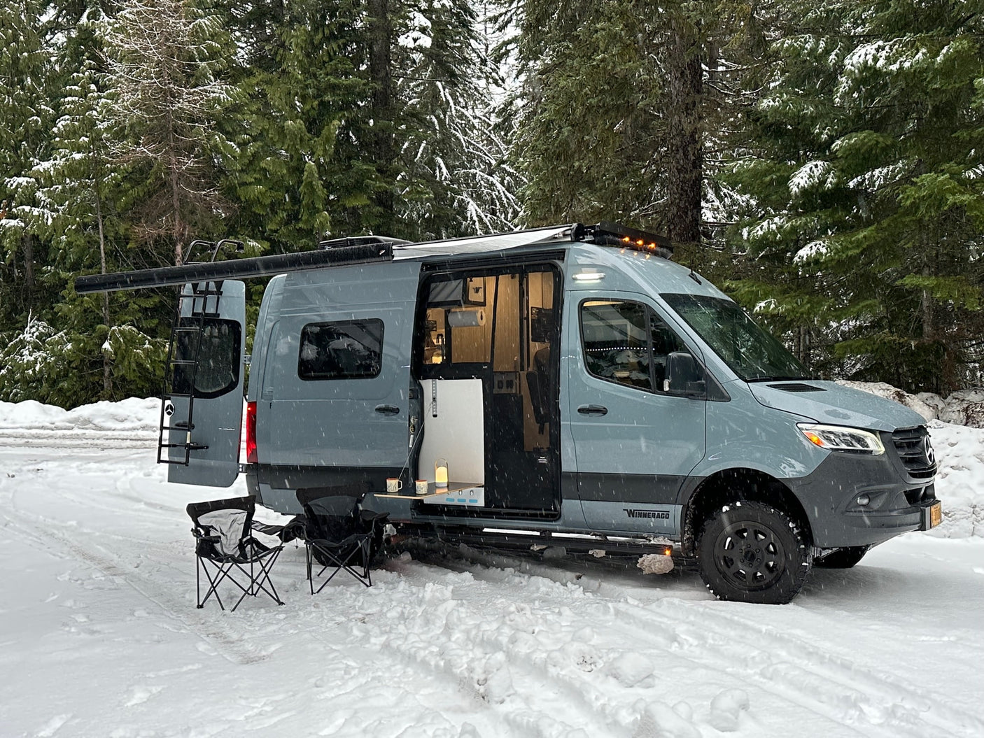 A sleek camper van, ideal for an rv rental, is parked in a snow-covered forest with its side door open, showing a cozy setup.