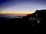 An RV rental parked at dawn, overlooking a sea of clouds and a vibrant blue-to-orange sky. Scenic mountain adventure awaits.