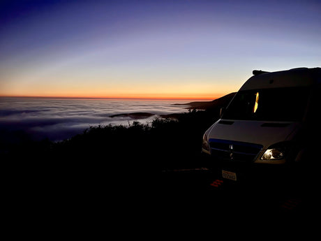 An RV rental parked at dawn, overlooking a sea of clouds and a vibrant blue-to-orange sky. Scenic mountain adventure awaits.