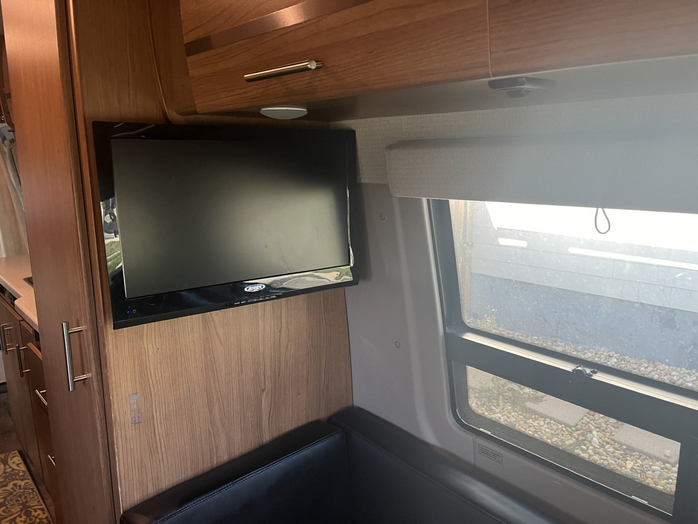 Inside an RV rental: view of mounted TV, wooden cabinets, black dinette cushion, and window with outdoor gravel view. — photo 14
