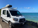 White Ford Transit RV rental parked oceanfront under a bright blue sky. Equipped with a roof rack and cargo box for travel.