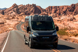 A sleek black Ram ProMaster RV rental drives on a winding desert road, set against a stunning backdrop of red rock mountains.