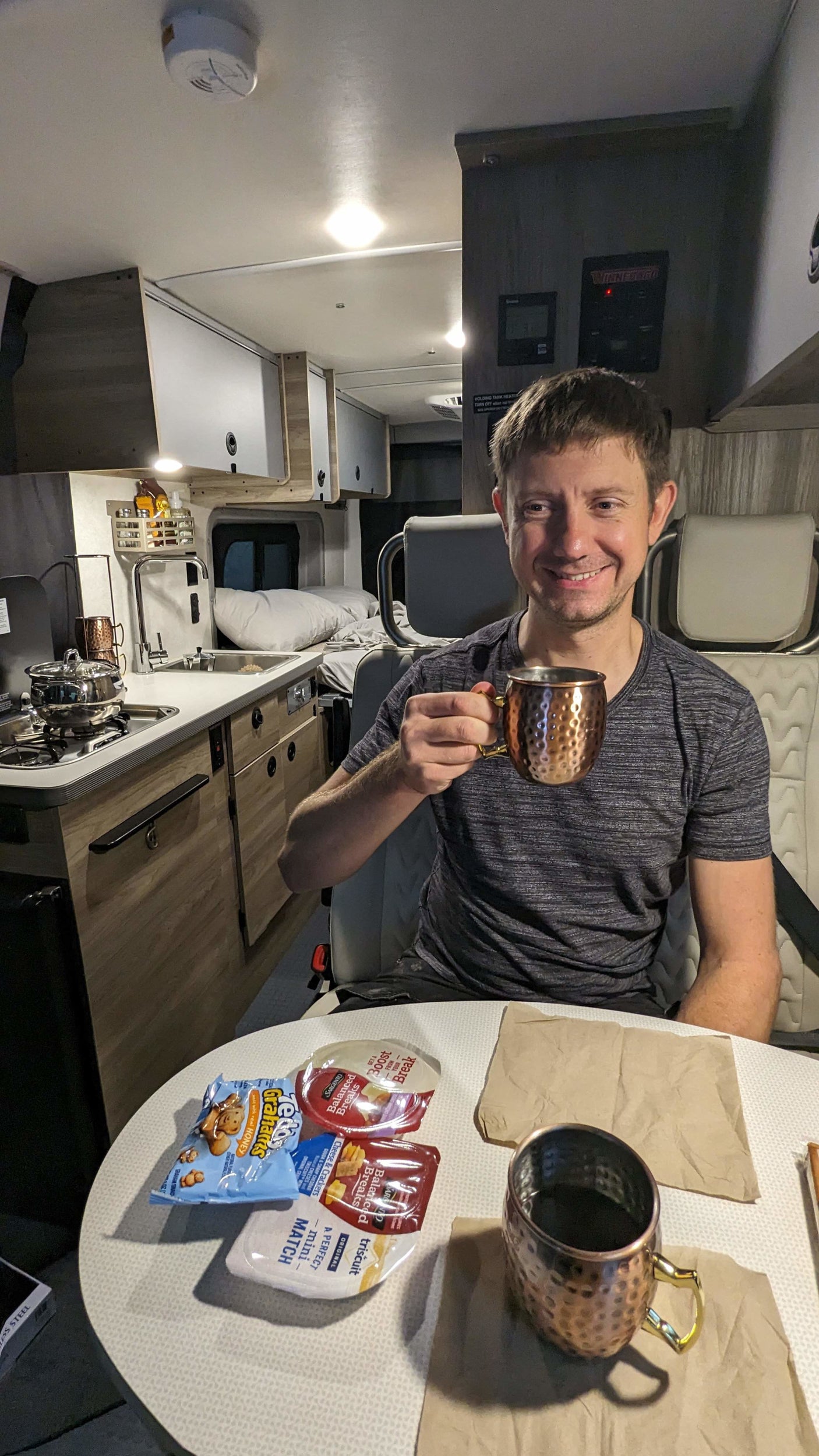 Smiling man holds a mug in a cozy RV rental. Snacks and a compact kitchen are visible, perfect for mobile travel.