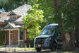 A dark Mercedes-Benz Sprinter RV rental parked before a historic brick house with a shingled roof, surrounded by lush green trees.