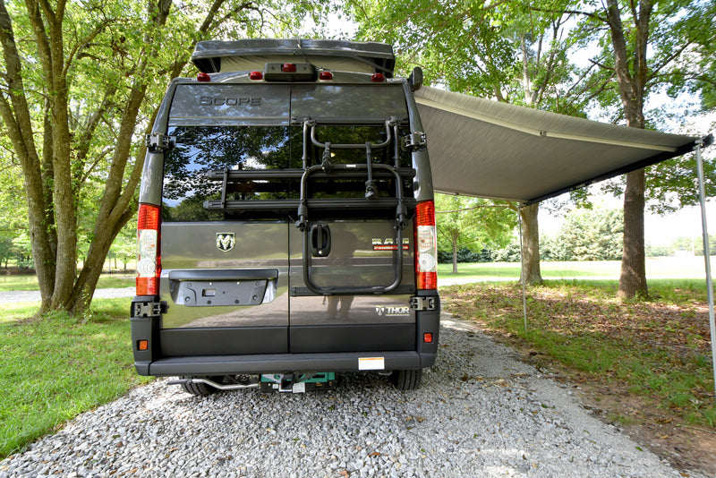 Rear of a Thor Scope camper van RV rental, featuring a bike rack and extended awning. Parked on gravel, nestled among green trees. — photo 16