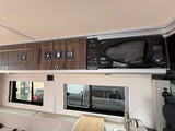 View of an RV rental kitchen with dark wood overhead cabinets, a microwave, and control panel above the sink and windows.