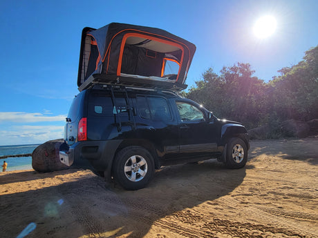 Explore off-grid with this black SUV featuring a rooftop tent, perfect for a unique rv rental experience on a sunny beach.