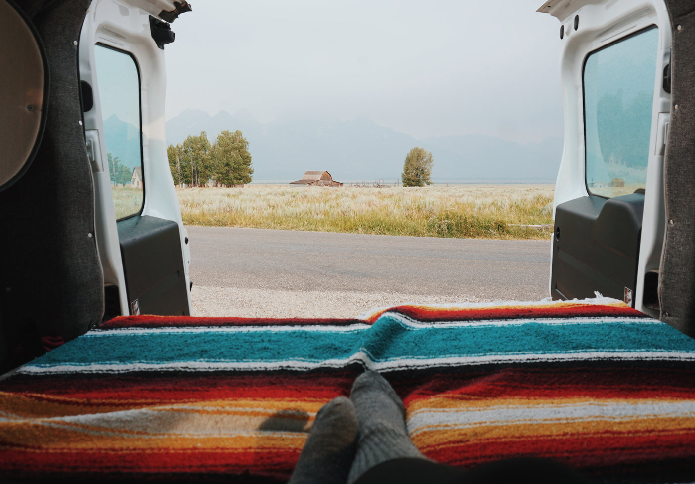 Cozy RV rental view: feet on a colorful blanket, gazing out at a serene field, historic barn, and misty mountains.