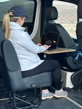 A woman in a grey hoodie & cap uses a laptop on a pull-out table inside an RV rental, viewing desert scenery.
