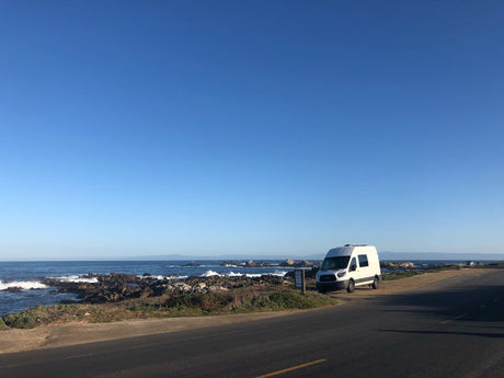 Coastal views with an RV rental! White camper van by the road, waves crashing on rocks, distant mountains under blue sky.