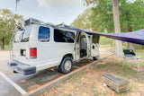 White camper RV rental with side awning, portable stove, and grill ready for outdoor cooking in a nature setting.