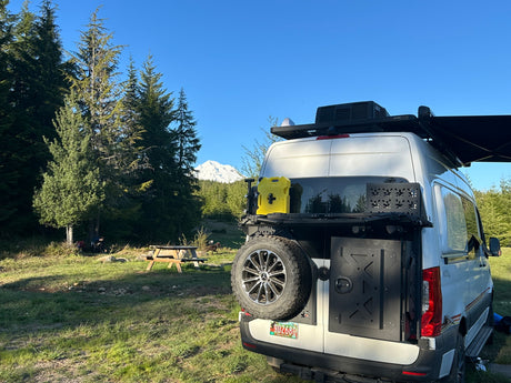 An RV rental campervan at a stunning mountain campsite, surrounded by tall pines, a snow-capped peak, and a picnic table.