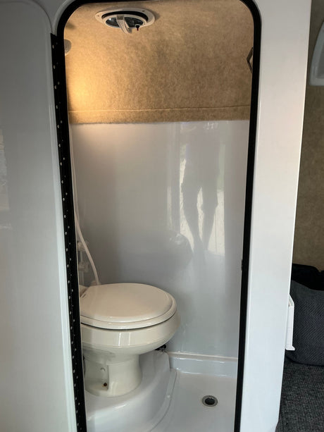 Compact bathroom in an RV rental, featuring a toilet, integrated shower pan, and ceiling vent. A person's reflection is visible.
