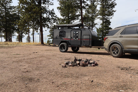 A gray teardrop camper trailer towed by an SUV at a rustic campsite with a fire pit, perfect for an rv rental outdoor adventure.