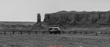 An RV rental camper van is parked on a dirt lot, foregrounded by a fence, against towering desert rock formations.
