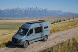 A light blue Mercedes Sprinter camper van on a dirt road, with panoramic mountain range views. Ideal for an RV rental trip.