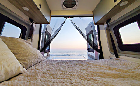 Cozy RV rental bed overlooks a tranquil beach and ocean at dawn/dusk through the open van doors.
