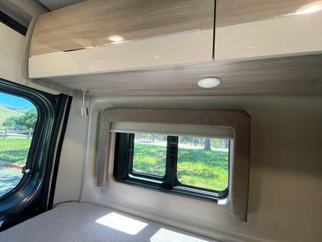 Inside an RV rental: a light-colored bed beneath a window revealing a green outdoor scene, with modern overhead cabinets and lighting.