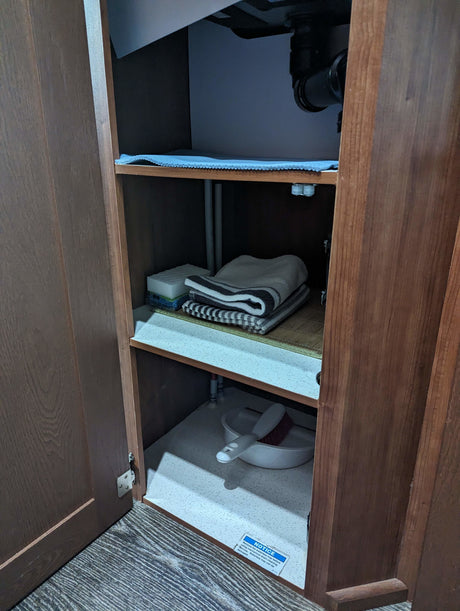 RV rental under-sink storage cabinet. Features plumbing, notice for a waterless trap, towels, sponges, & cleaning items.
