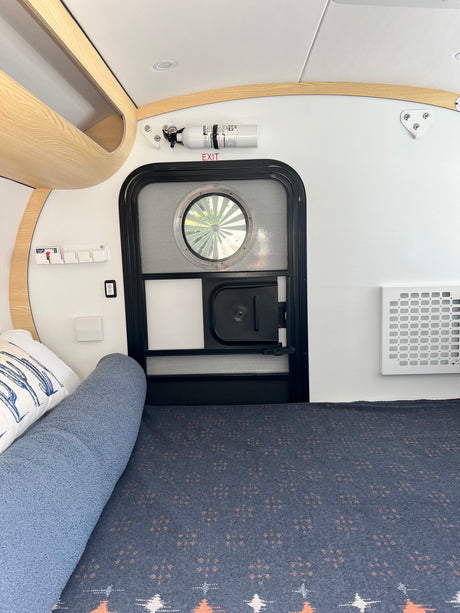 Interior of a compact rv rental, showing a bed, main entry door with circular window, and fire extinguisher mounted above.