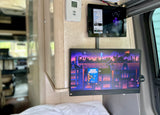 RV rental interior: Two wall-mounted screens with a media library tablet and a monitor showing a college basketball ad; thermostat visible.