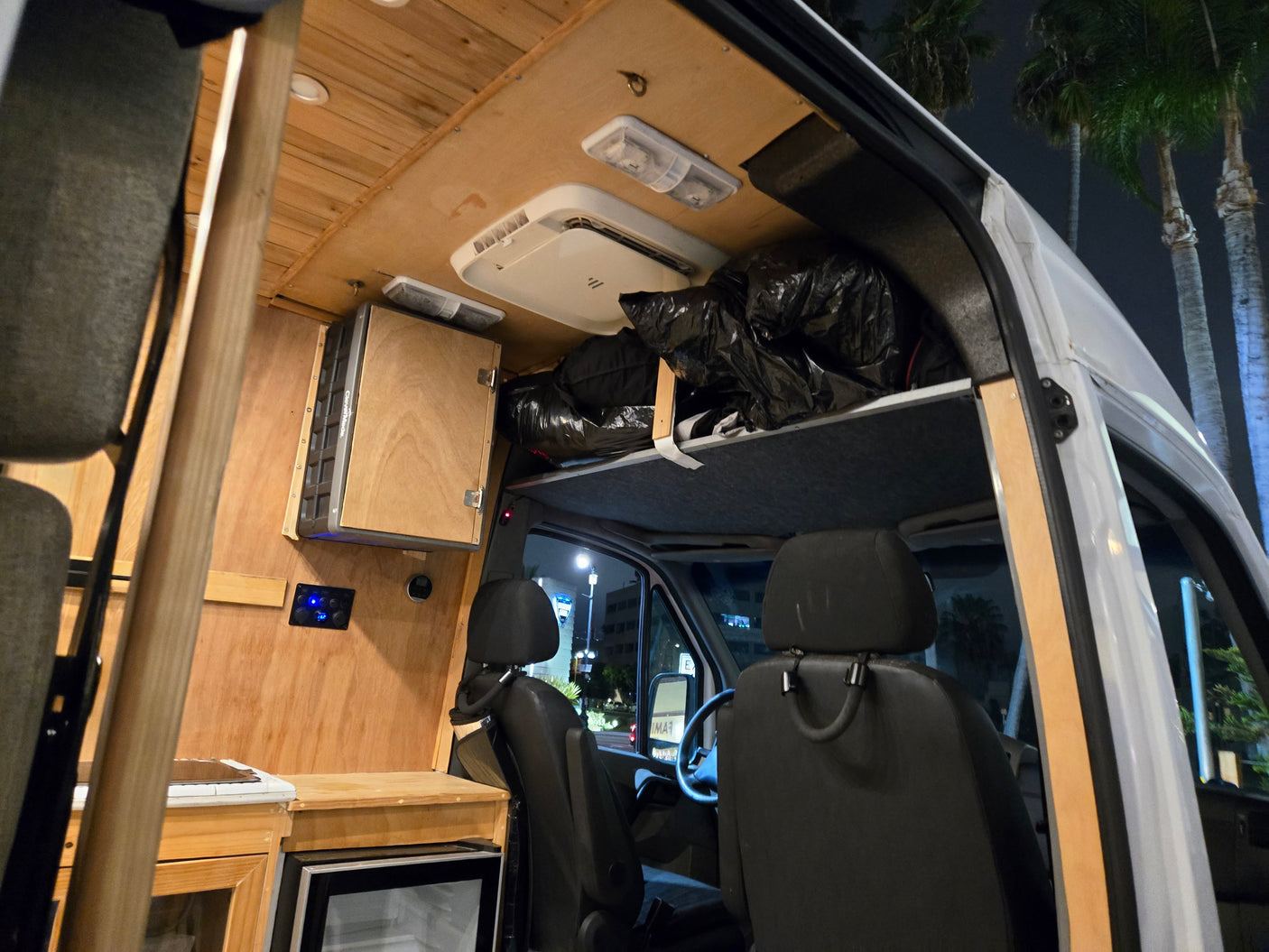 Inside a cozy RV rental van: wooden interior, control panel, fridge, and overhead storage, illuminated at night.