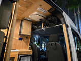 Inside a cozy RV rental van: wooden interior, control panel, fridge, and overhead storage, illuminated at night.