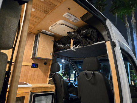 Inside a cozy RV rental van: wooden interior, control panel, fridge, and overhead storage, illuminated at night.