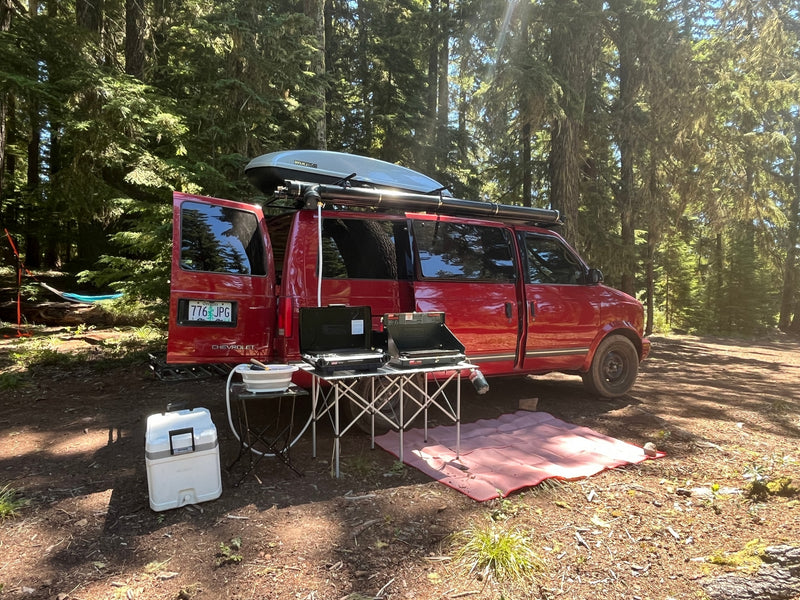 Red Chevy van, set up as an RV rental, camped in a sunny forest clearing with outdoor kitchen, hammock, and gear. — photo 2