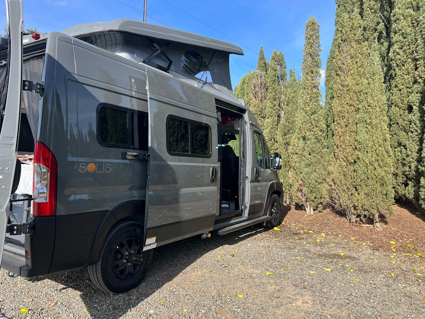 Gray Winnebago Solis pop-top camper van RV rental parked on gravel near trees, doors open.