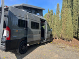 Gray Winnebago Solis pop-top camper van RV rental parked on gravel near trees, doors open.