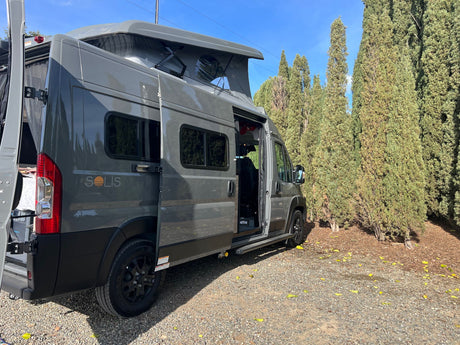 Gray Winnebago Solis pop-top camper van RV rental parked on gravel near trees, doors open.