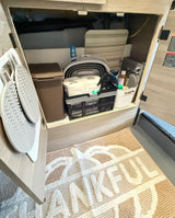 RV rental storage: cabinet with TP, cleaning supplies, and collapsible bin. Mats hang on door, "THANKFUL" rug below.