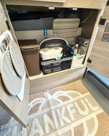 RV rental storage: cabinet with TP, cleaning supplies, and collapsible bin. Mats hang on door, "THANKFUL" rug below.