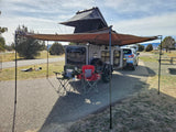 Campsite ready: Grey camper trailer with rooftop tent & awning, SUV, chairs, bike. Great for RV rental adventures!