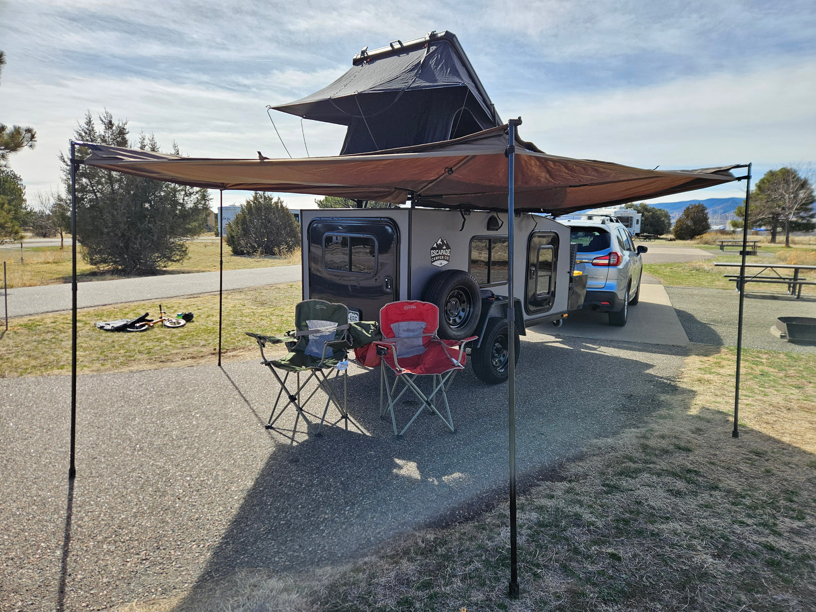 Campsite ready: Grey camper trailer with rooftop tent & awning, SUV, chairs, bike. Great for RV rental adventures!