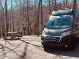Dark grey RV rental parked on a gravel campsite with a picnic table & grill, surrounded by bare trees.