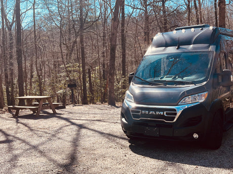 Dark grey RV rental parked on a gravel campsite with a picnic table & grill, surrounded by bare trees.