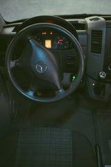 Driver's perspective inside a Mercedes-Benz Sprinter van for RV rental, showing the steering wheel, lit dashboard, & gear shifter.