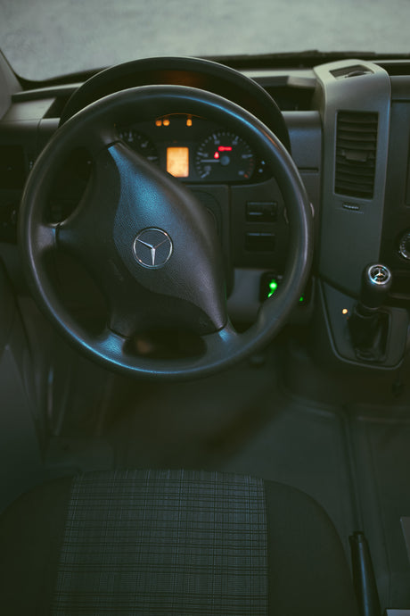 Driver's perspective inside a Mercedes-Benz Sprinter van for RV rental, showing the steering wheel, lit dashboard, & gear shifter.