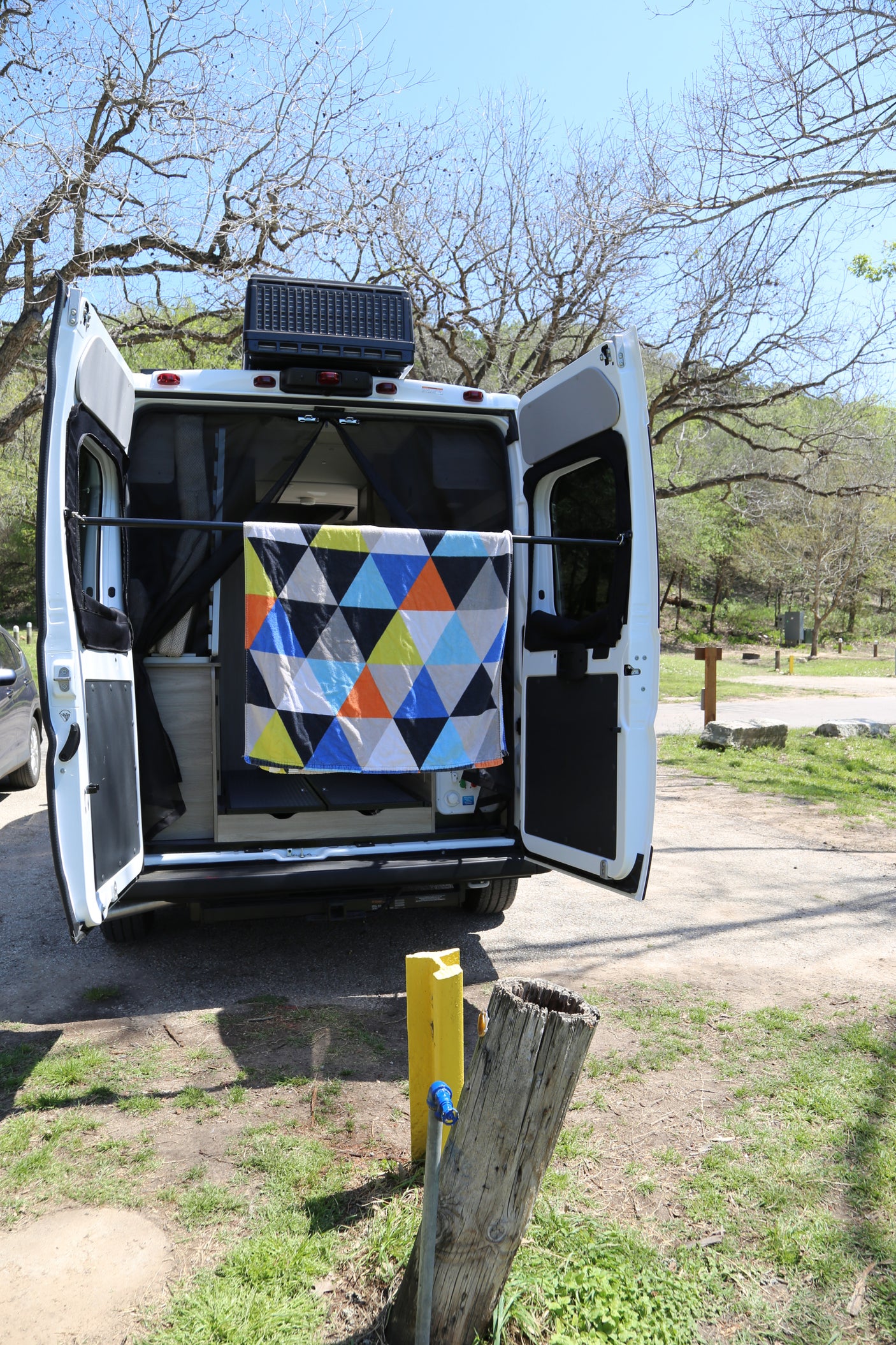 RV rental camper van: Back doors open, a colorful blanket dries in the sun at a scenic campsite. Water spigot near.