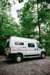 White Winnebago Solis Pocket camper van for rv rental on gravel, surrounded by lush green forest. Perfect for nature travel.