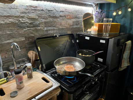 This compact RV rental kitchen features a gas stove cooking a meal, with a sink and spices ready for adventure.