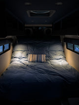 This RV rental's cozy bed features denim-blue bedding, a striped pillow, and bright reading lights for comfortable travels.